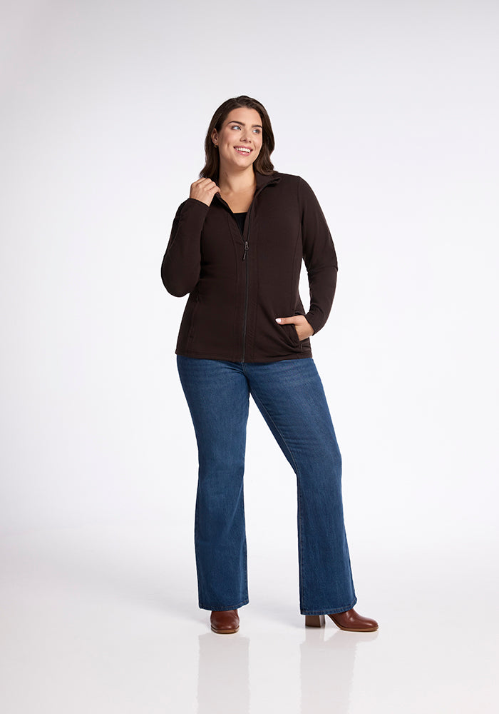 A person is standing against a plain background, smiling and looking off to the side. They are wearing a sleek French Roast Finley Zip Up Sweatshirt from Woolx, blue bell-bottom jeans, and brown boots. One hand is in their pocket while the other hand is touching their hair.