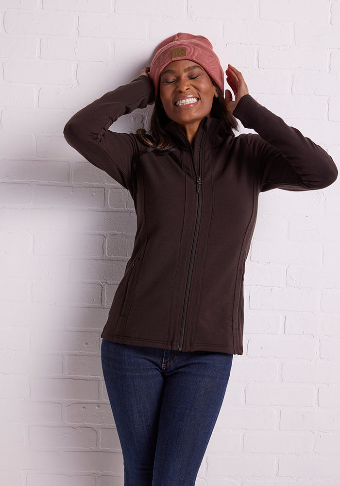 A woman stands against a white brick wall, smiling with her eyes closed and pulling on her Brick Dust Baylor Beanie from Woolx. She is wearing a dark zip-up jacket and blue jeans.