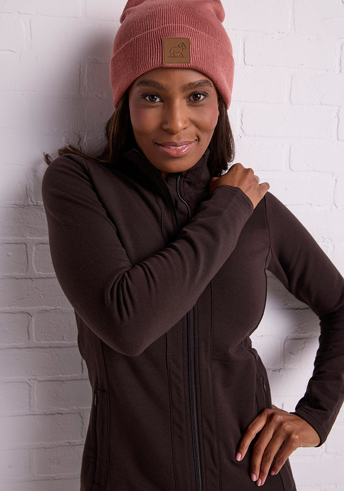 A person is posing in front of a white brick wall. They are wearing a dark brown zip-up jacket and the Woolx Baylor Beanie in Brick Dust, made from warm Merino wool. They are smiling and have one hand resting on their opposite shoulder.