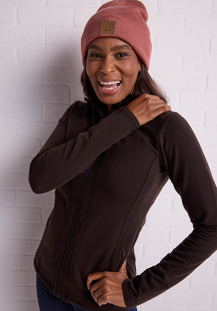 A person is smiling while wearing a brown zip-up jacket and the Baylor Beanie from Woolx, a pink hat adorned with a brown patch on the front. The background consists of a white brick wall. The cozy look is completed with what seems to be this warm winter hat, perfect for chilly days.