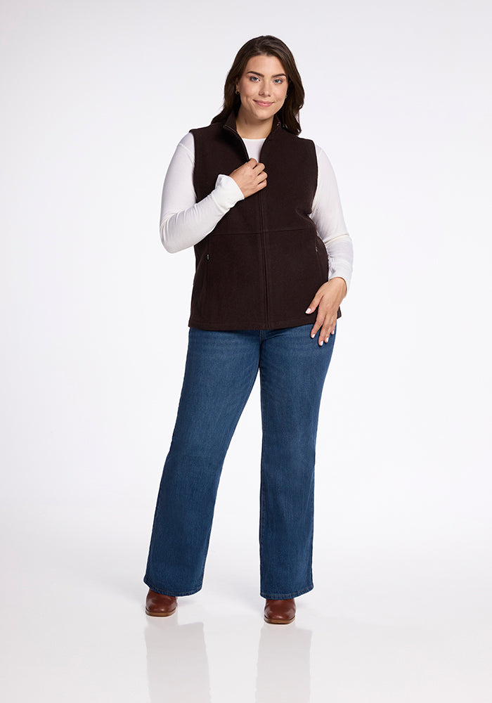 A woman with shoulder-length dark hair is wearing the Colbie Vest in French Roast by Woolx over a white long-sleeve shirt, blue jeans, and brown shoes. She is standing with one hand touching the collar of her vest and the other resting on her hip, posing against a plain white background.