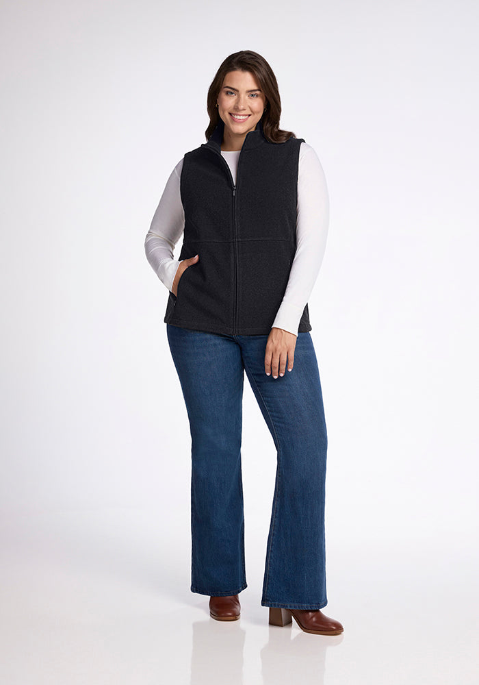 A smiling woman stands against a plain backdrop, wearing the Woolx Colbie Vest in Carbon Black over a white long-sleeve shirt, paired with blue jeans and brown boots. She has one hand in her pocket and the other resting by her side.