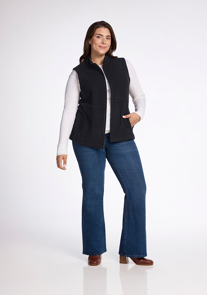 A person stands confidently against a plain white background, donning the Woolx Colbie Vest in Carbon Black over a white long-sleeve shirt. They pair this stylish ensemble with blue jeans that flare at the bottom and brown shoes. With one hand resting in the pocket of the vest, they flash a cheerful smile.