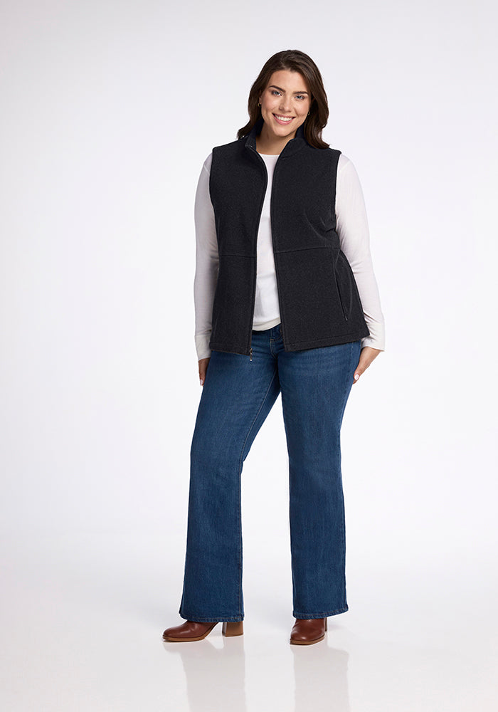 A smiling person stands against a plain white background. They are wearing a Colbie Vest in Carbon Black by Woolx over a white long-sleeve shirt, blue jeans, and brown shoes. They have one hand in a pocket and are in a relaxed, confident stance.