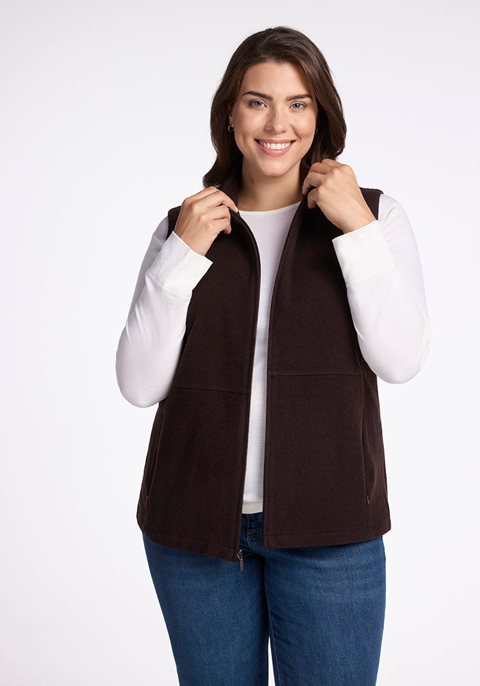 A woman with long brown hair is smiling while wearing the Woolx Colbie Vest in French Roast over a white long-sleeved shirt and blue jeans. She is holding the collar of her vest with both hands, against a plain white background. 