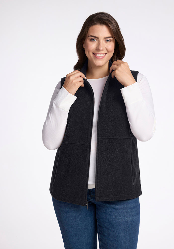 A woman with shoulder-length brown hair is smiling at the camera. She is wearing a Woolx Colbie Vest in Carbon Black over a white long-sleeved shirt and blue jeans. She is holding the collar of the vest with both hands. The background is plain white. 