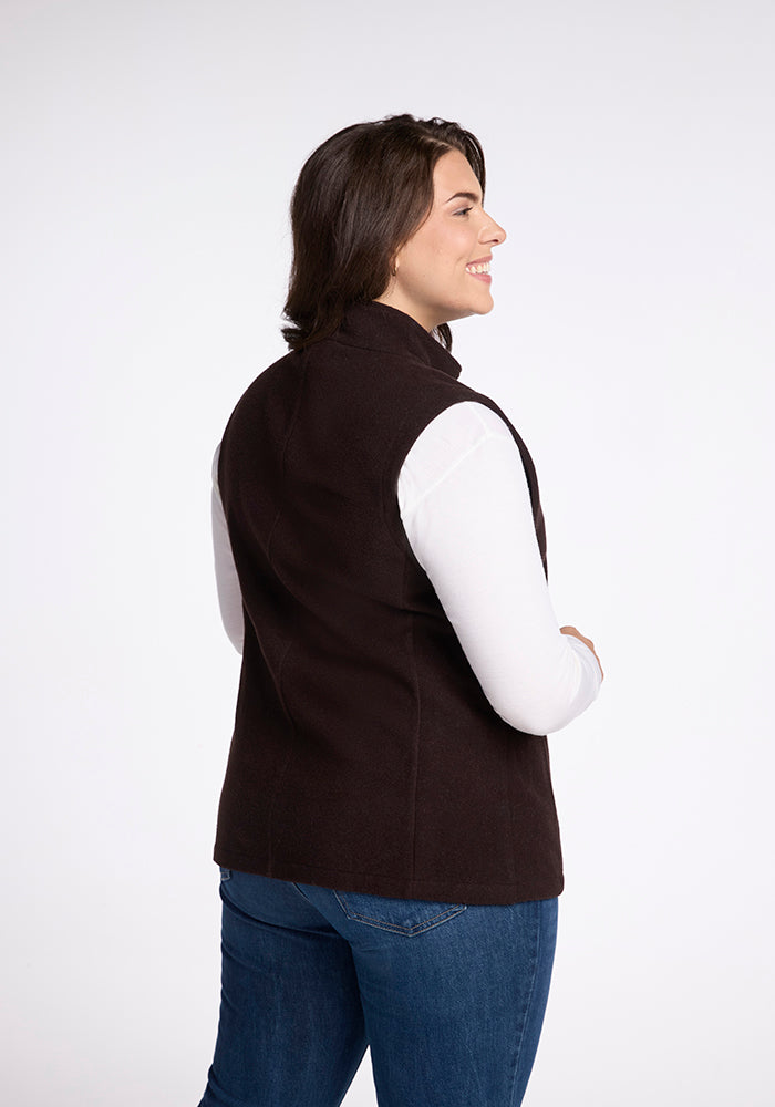 A woman with long dark hair is shown from the back, smiling and looking over her right shoulder. She is wearing the Woolx Colbie Vest in French Roast over a white long-sleeved shirt and blue jeans. The background is plain white.