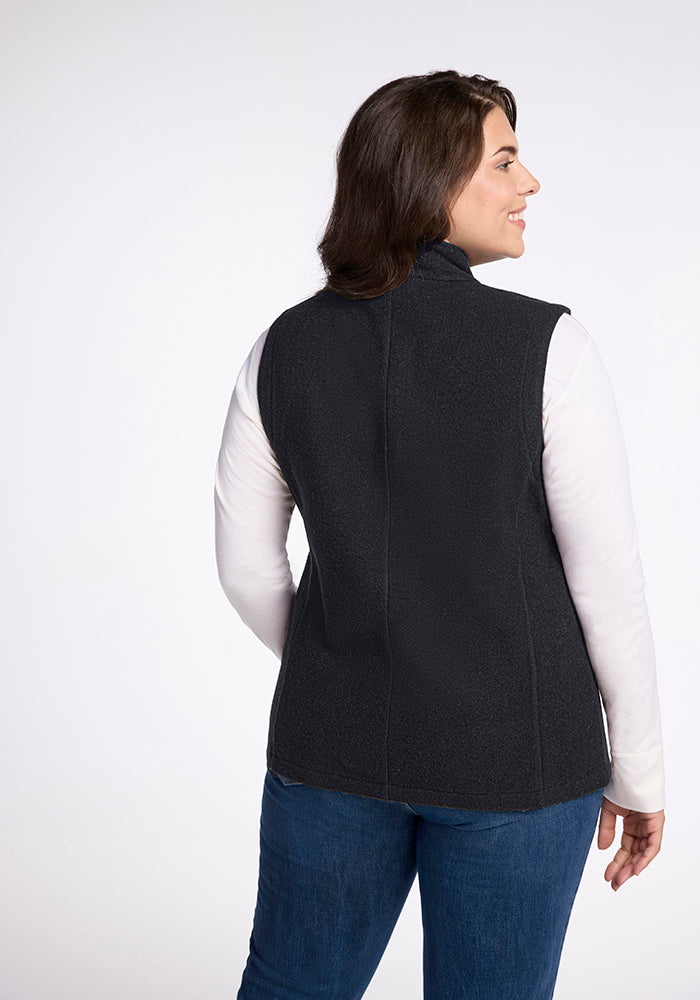 A woman with brown hair is facing away from the camera, sporting the Colbie Vest - Carbon Black by Woolx over a white long-sleeve shirt. She pairs this with blue jeans and has a slight smile, standing against a white background.