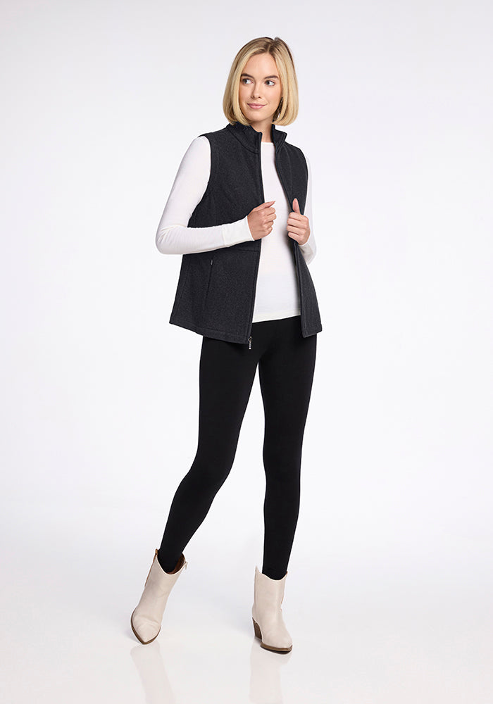 A woman with shoulder-length blonde hair is standing and facing forward. She is wearing a Woolx Colbie Vest in Carbon Black over a white long-sleeve shirt, black pants, and light-colored ankle boots. She has a slight smile and is holding the edges of her vest with both hands.