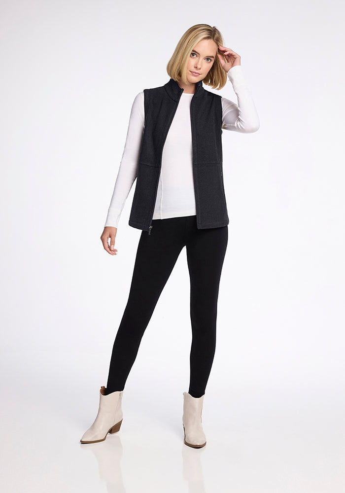 A woman with shoulder-length blond hair is wearing the Woolx Colbie Vest in Carbon Black over a white long-sleeve top, paired with black pants and white ankle boots. She is standing against a plain white background, resting one hand on her head and looking at the camera.