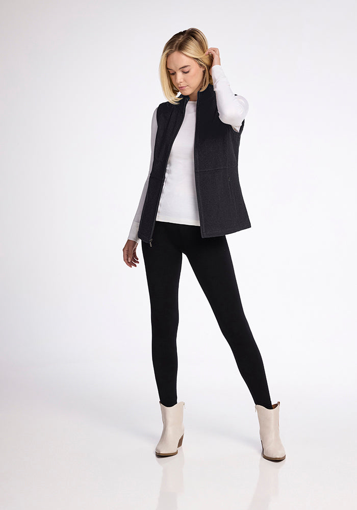 A woman stands confidently in front of a plain background, wearing a white long-sleeve top, the Colbie Vest in Carbon Black from Woolx, black leggings, and white ankle boots. She has blonde hair and is looking downwards with one hand placed near her neck.