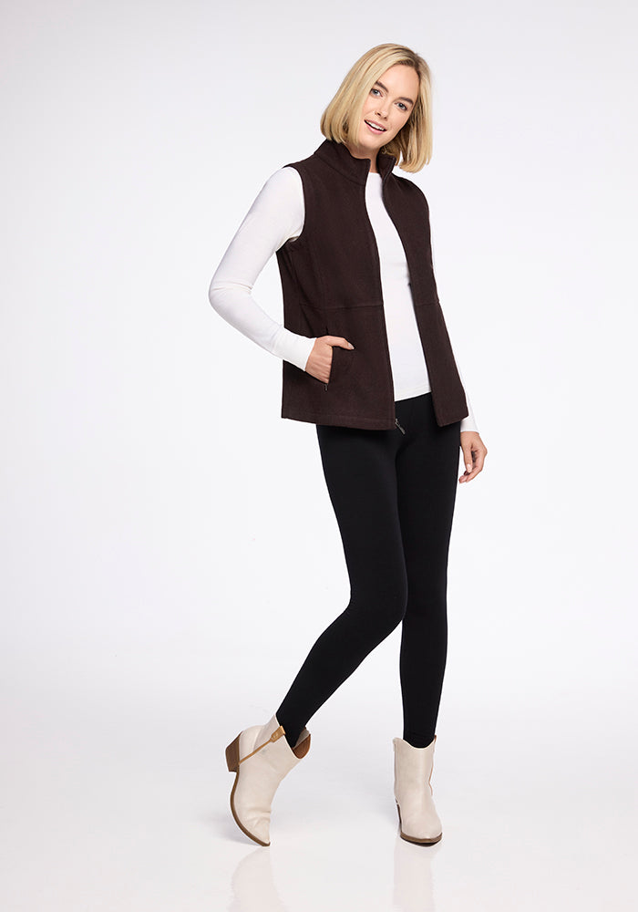 A woman with shoulder-length blonde hair is posing against a plain white background. She is wearing a white long-sleeved shirt, the Colbie Vest - French Roast by Woolx, black leggings, and beige ankle boots. She is smiling and has one hand in her vest pocket.