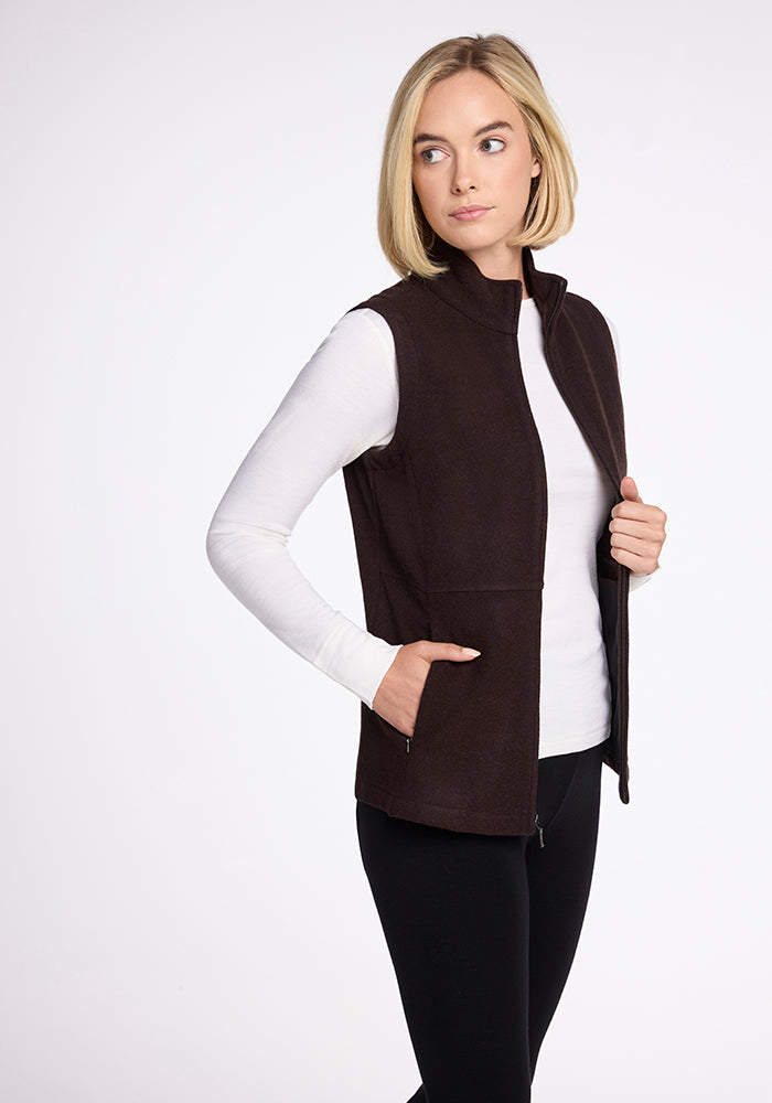 A woman with shoulder-length blonde hair is wearing a Woolx Colbie Vest in French Roast over a white long-sleeve shirt and black pants. She is facing slightly to the side with one hand in her pocket and the other adjusting the zipper. The background is plain white.