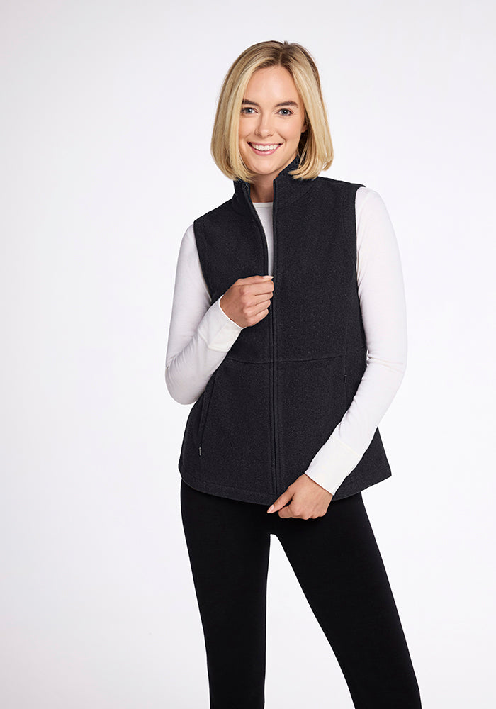 A smiling person with shoulder-length blonde hair is wearing a white long-sleeve shirt under the Woolx Colbie Vest in Carbon Black. They are standing against a plain white background and holding the vest with one hand near the zipper.