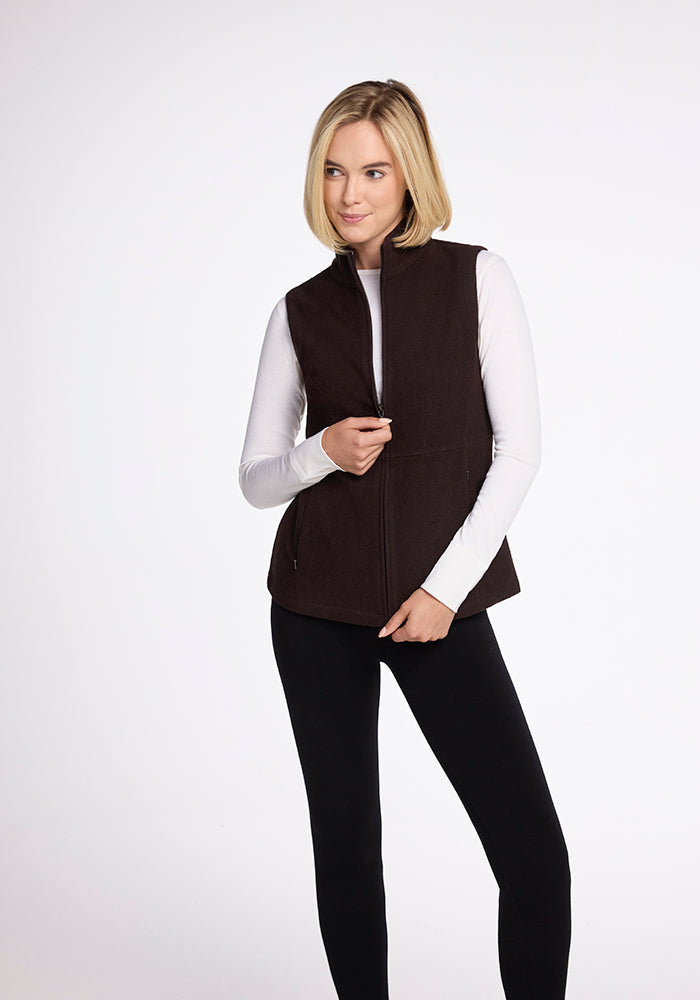 A person with shoulder-length blonde hair wearing the Woolx Colbie Vest in French Roast over a white long-sleeve shirt and black pants stands against a plain white background. Their left hand is touching the zipper of the vest while looking off to the side.