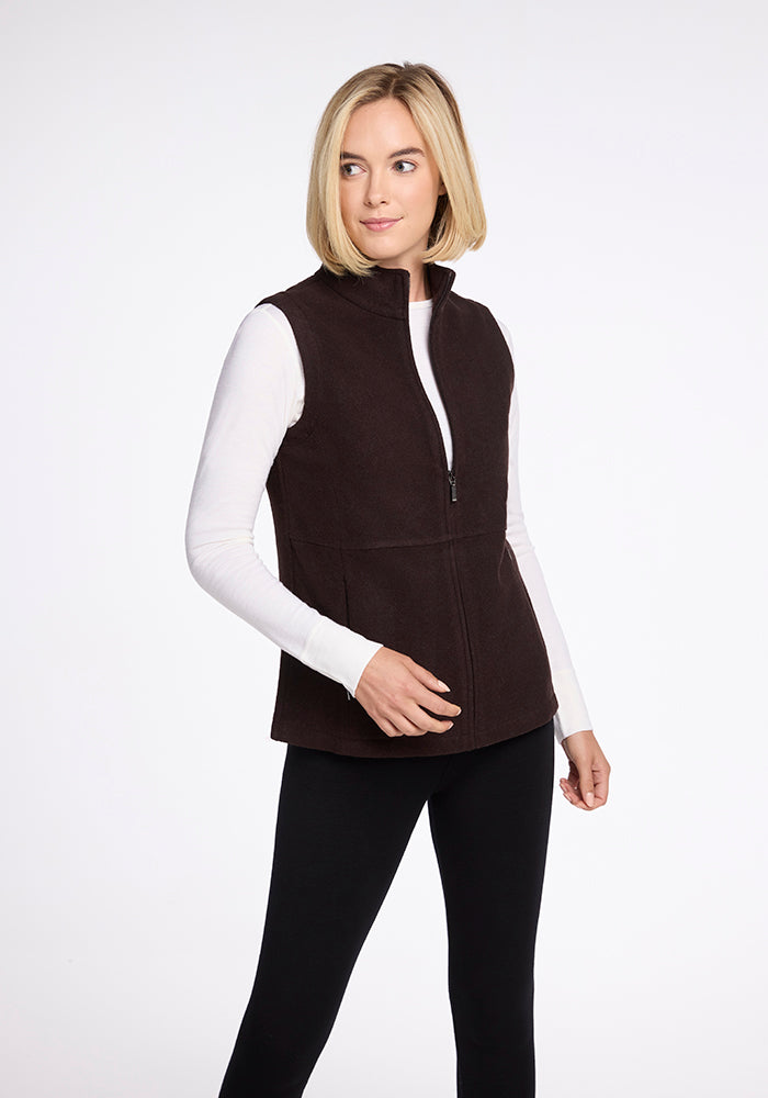 A woman with blonde hair is wearing a French Roast Colbie Vest by Woolx over a white long-sleeve shirt and black pants. She stands against a plain white background, smiling slightly and looking to her right. 