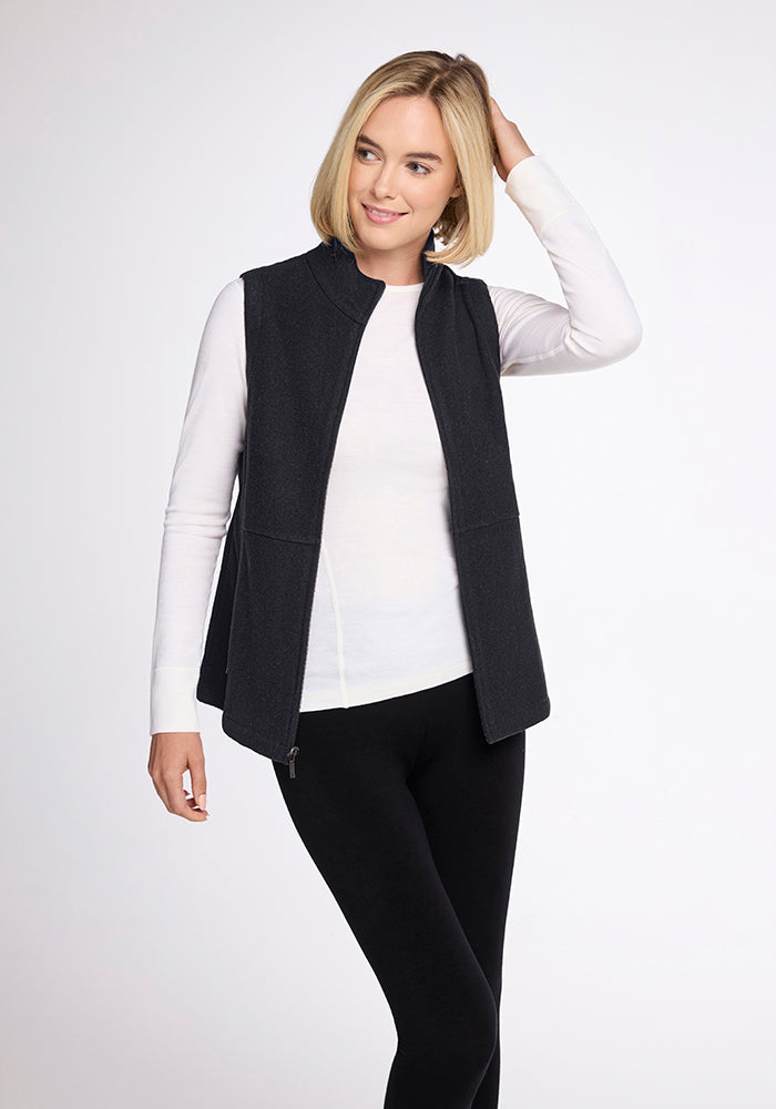 A woman with shoulder-length blonde hair wears a Woolx Colbie Vest in Carbon Black over a long-sleeve white shirt and black leggings. She stands against a plain light-colored background, smiling and raising her left hand to touch her hair.