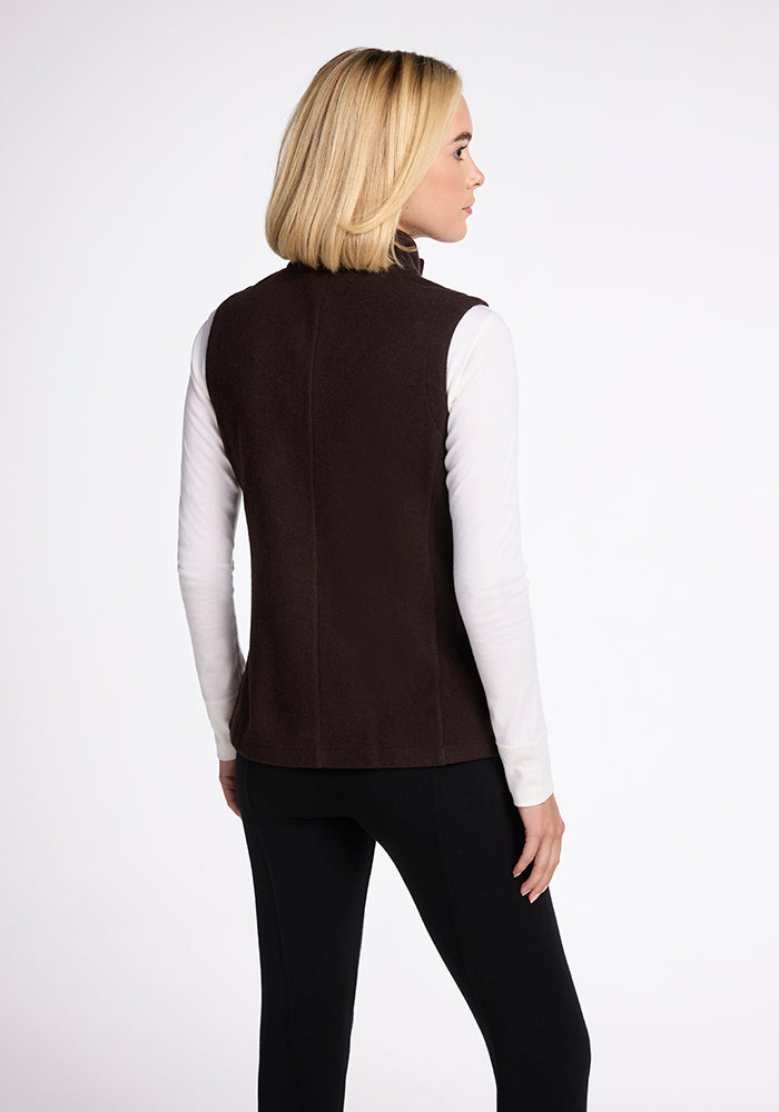 A person with shoulder-length blonde hair is seen from the back, wearing the Woolx Colbie Vest in French Roast over a long-sleeved white top and black pants. The background is plain and light-colored.