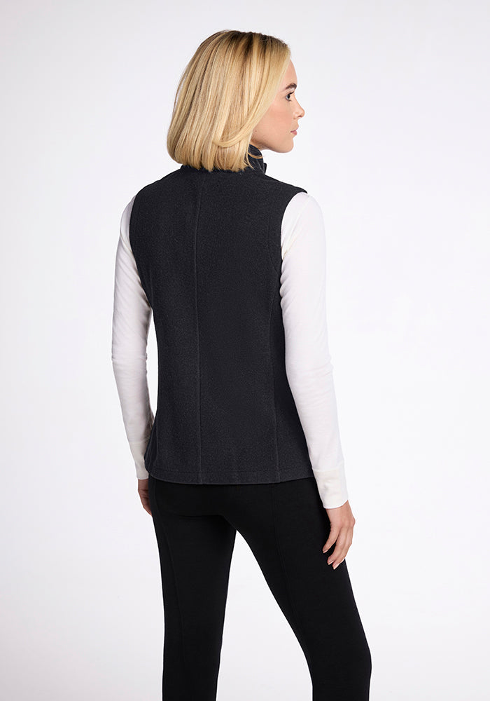 A person with shoulder-length blonde hair is standing and facing away from the camera. They are wearing the Colbie Vest in Carbon Black by Woolx over a long-sleeved white shirt, paired with black pants. The background is plain light grey.