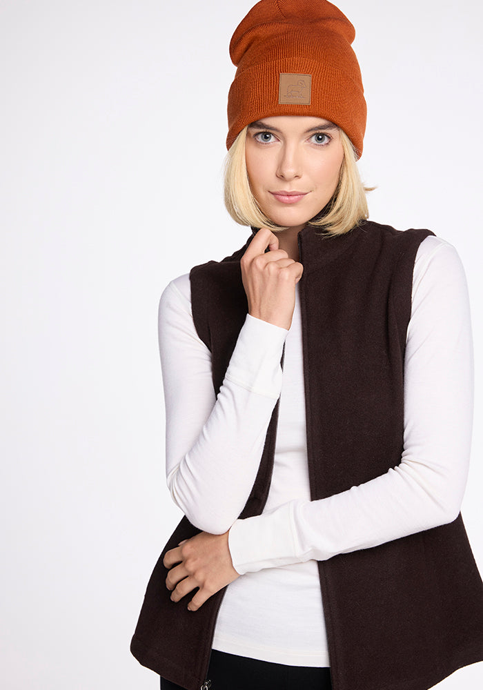 A woman with blonde hair is wearing a Woolx Baylor Beanie - Cinnamon Stick, a white long-sleeve shirt, and a dark brown vest. She is looking at the camera with a slight smile and has one hand gently touching her chin. The background is plain white.