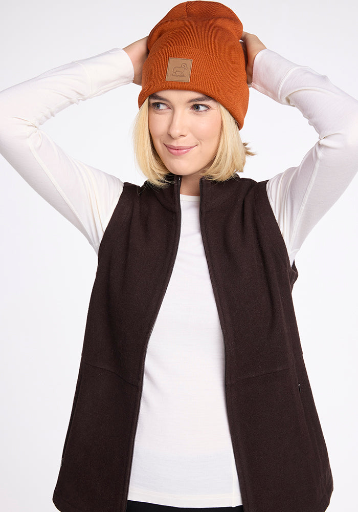 A person with shoulder-length blonde hair is adjusting a Woolx Baylor Beanie on their head. They are wearing a white long-sleeve shirt beneath a sleeveless, dark brown vest. The background is plain white.