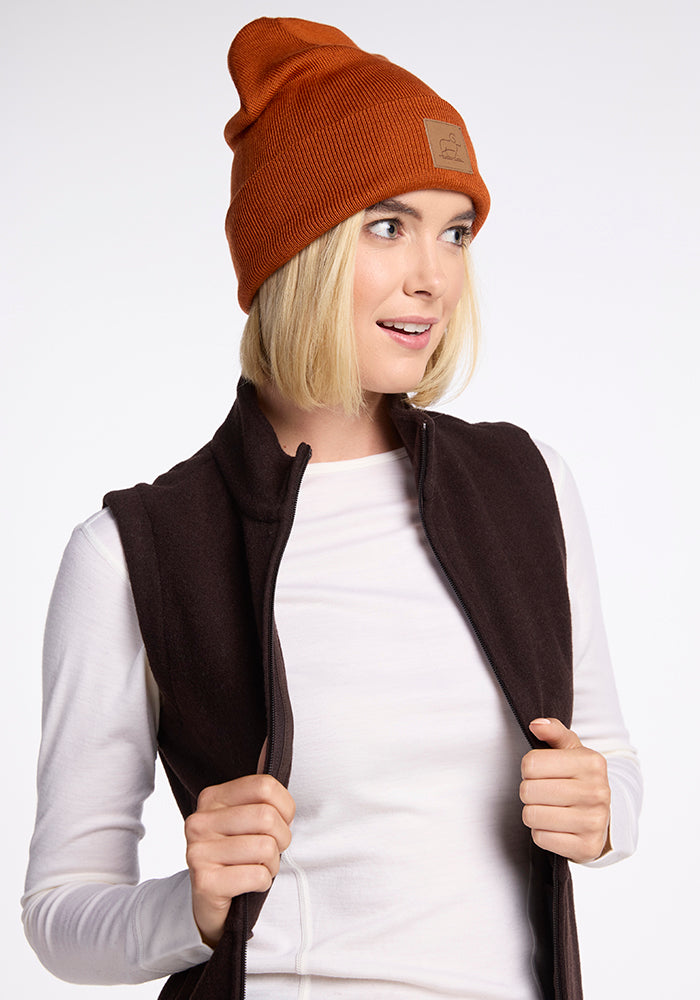 A person with a short blonde bob hairstyle is wearing an orange Woolx Baylor Beanie, a white long-sleeve shirt, and a brown zip-up vest. They are looking to the side and holding the edges of the vest with both hands. The background is plain and well-lit.