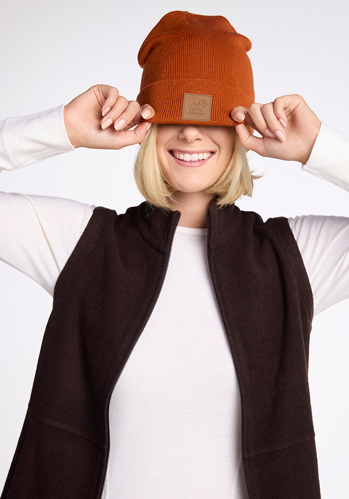 A blonde woman is smiling as she playfully pulls a Woolx Baylor Beanie in Cinnamon Stick down over her eyes. She is dressed in a white long-sleeve shirt under a dark brown vest, set against a plain, light-colored background that highlights her cheerful demeanor.