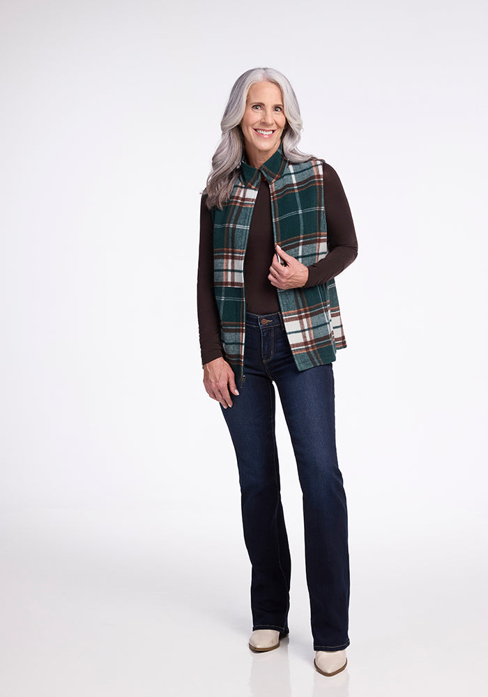 A woman with long gray hair stands confidently in front of a plain white background. She is wearing the Woolx Colbie Vest in Espresso Plaid over a dark long-sleeved shirt, paired with dark blue jeans and beige shoes. She smiles and has one hand holding the vest near her collar.