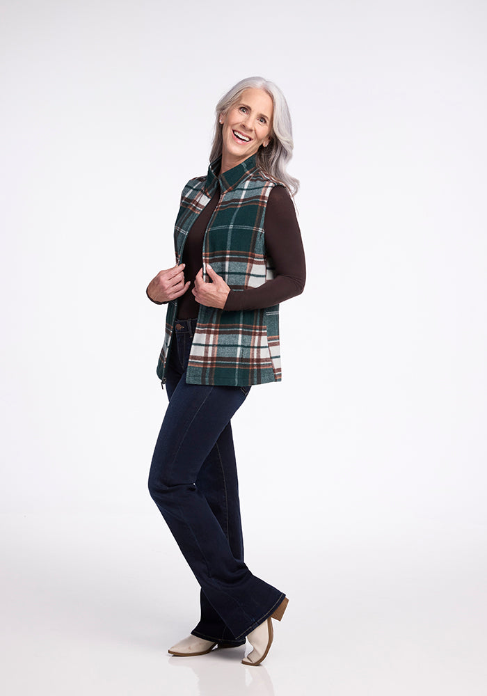 A woman with long gray hair smiles while posing in the Woolx Colbie Vest - Espresso Plaid over a dark long-sleeve shirt and dark jeans. She stands with one leg slightly bent, arms partially raised, against a plain white background. She wears light-colored shoes.