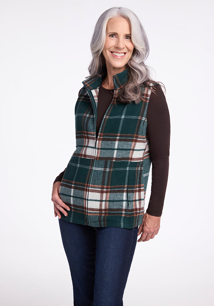 A smiling woman with long, wavy gray hair is wearing the Woolx Colbie Vest in Expresso Plaid over a dark long-sleeved shirt and blue jeans. She stands against a plain white background with one hand resting on her hip. 