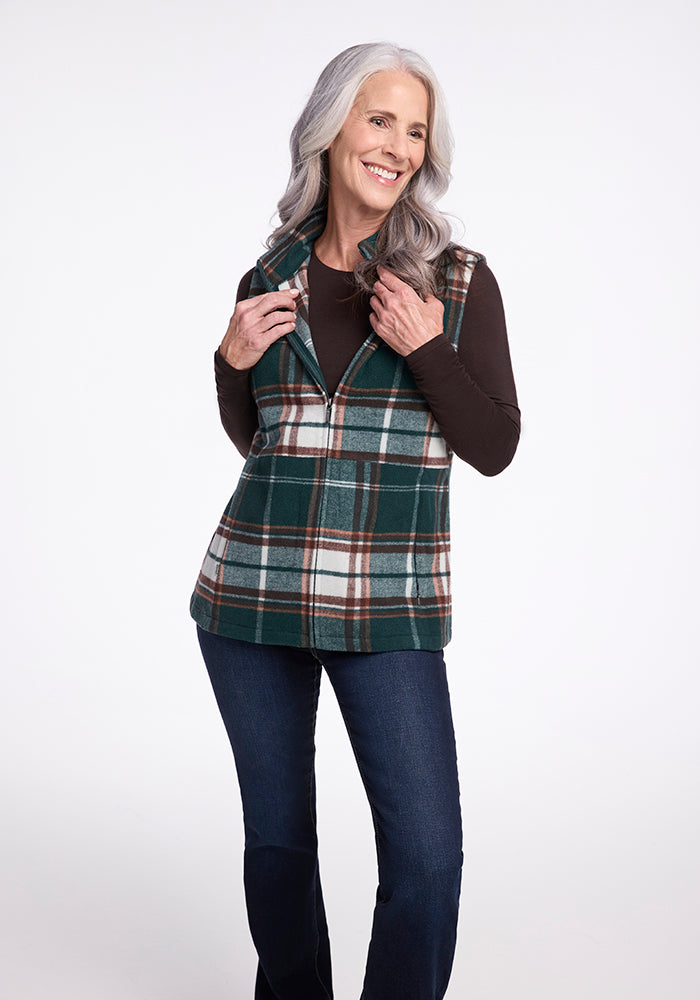 A woman with long gray hair smiles while posing in the Woolx Colbie Vest - Espresso Plaid, a sleeveless green, brown, and white plaid vest worn over a dark long-sleeve shirt. She is also wearing blue jeans and stands against a plain white background.