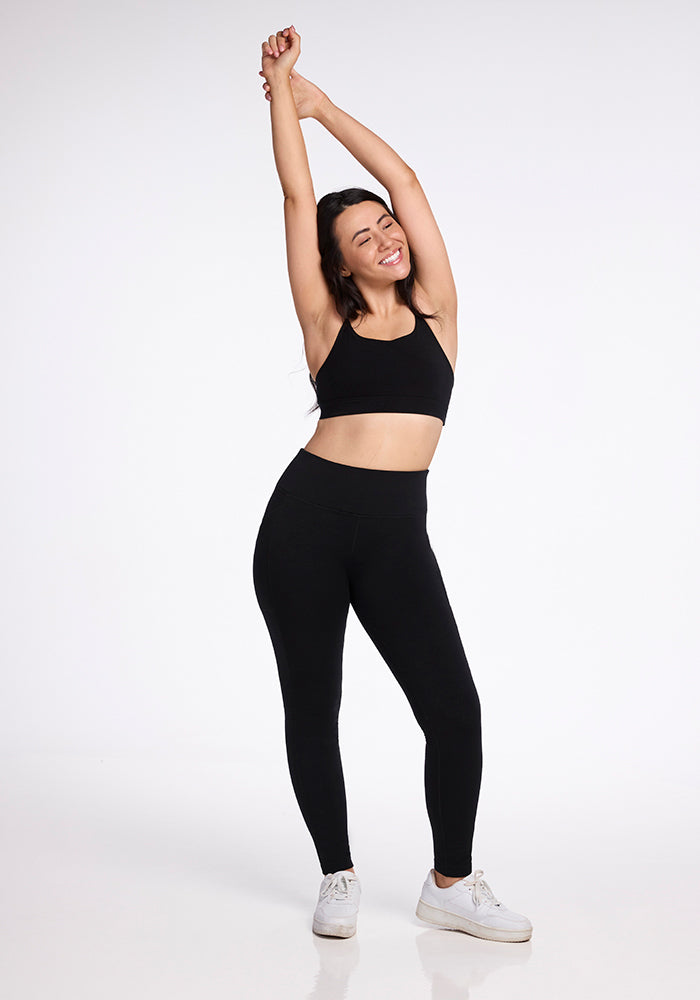 A woman stands with her arms stretched above her head, smiling and wearing a black sports bra, Frost Duralite® Leggings by Woolx in black, and white sneakers. The background is plain white.