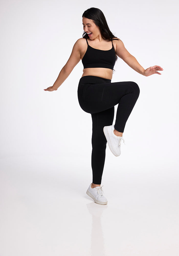 A woman wearing Woolx's Frost Duralite® Leggings in black, featuring convenient side pockets, and a pair of white sneakers is smiling as she balances on one leg with her arms outstretched. She is posing against a plain white background.