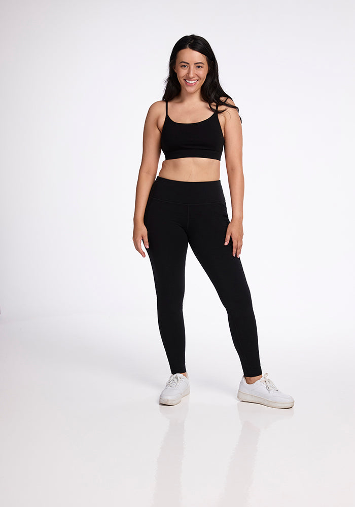 A woman with long dark hair is smiling and standing in a white space, wearing a black sports bra, Woolx Frost Duralite® Leggings - Black with side pockets, and white sneakers. She has her arms relaxed by her sides, and her reflection is faintly visible on the shiny floor.