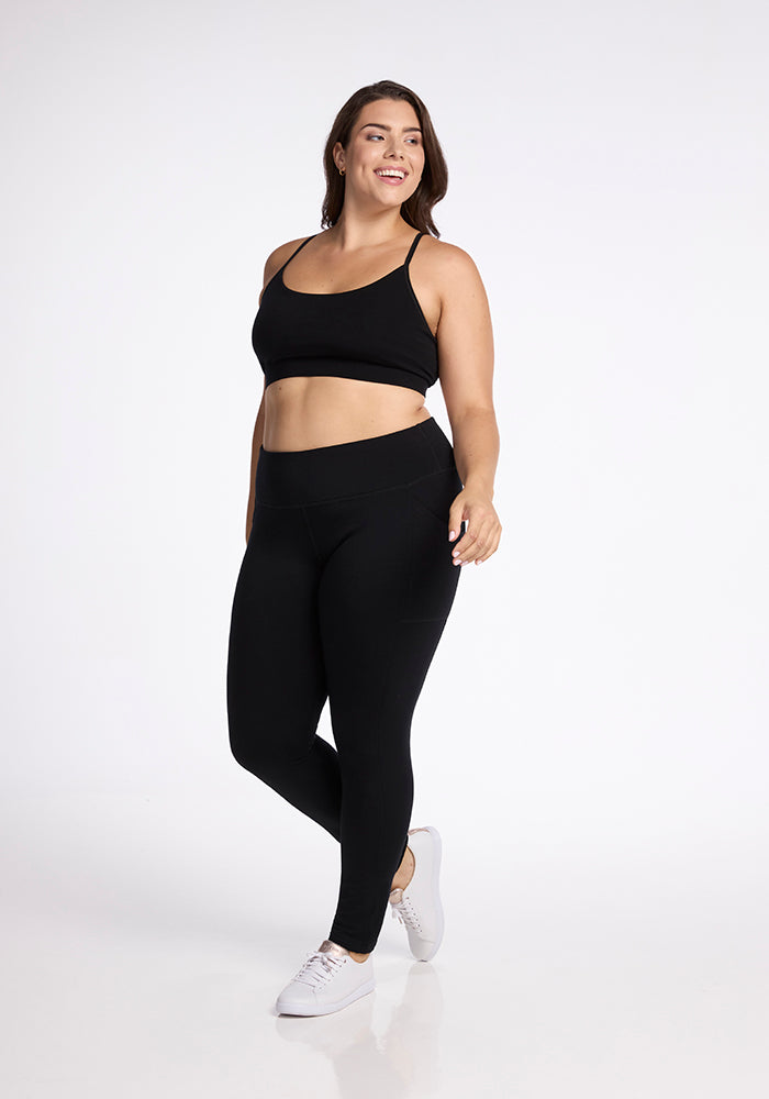 A woman poses confidently in Woolx's black Frost Duralite® Leggings and a matching sports bra against a simple white background. She has brown hair, wears white sneakers, and smiles while looking slightly to the side.