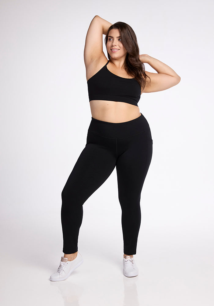 A woman with long brown hair poses confidently against a white background. She is dressed in a black sports bra, Woolx Frost Duralite® Leggings in Black, and white sneakers. Her left arm is bent with her hand resting on the back of her head, and her right arm is relaxed by her side.