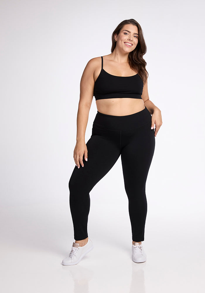 A woman poses confidently in Woolx's black Frost Duralite® Leggings and a matching sports bra, paired with white sneakers. She smiles and stands with one hand on her hip against a plain white background.