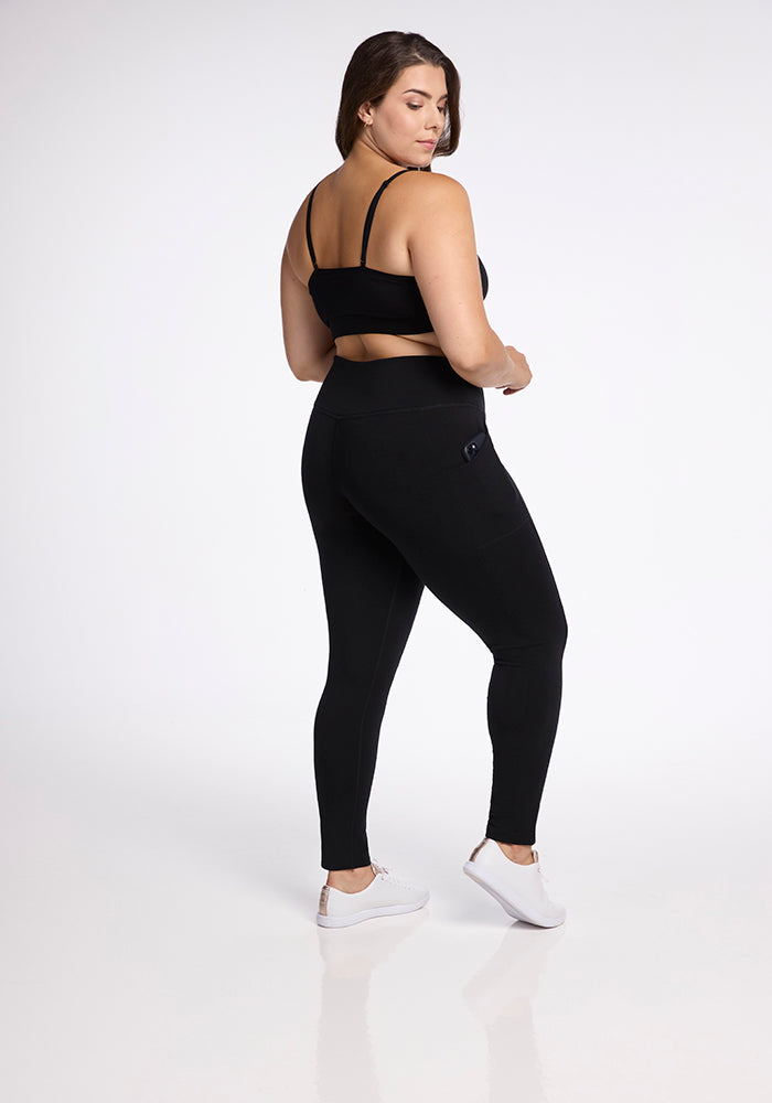 A woman stands facing away, slightly turning to her left. She is wearing a black sports bra, Frost Duralite® Leggings - Black by Woolx, and white sneakers. The background is plain white, highlighting her athletic outfit.