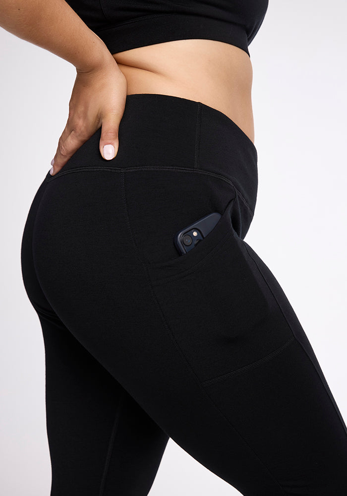 Close-up of a person's torso and upper leg, showing them wearing black Frost Duralite® Leggings from Woolx with a phone partially tucked into a side pocket. The person is placing their hand on their hip, and they are dressed in a matching black top.