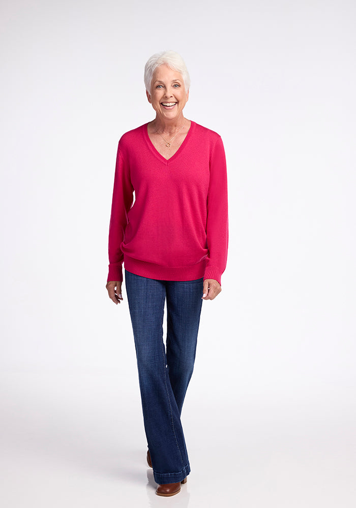 An older woman with short white hair is smiling and walking forward. She is wearing the Woolx Francesca Sweater in Berry Burst, along with blue jeans and brown shoes. The background is plain white.