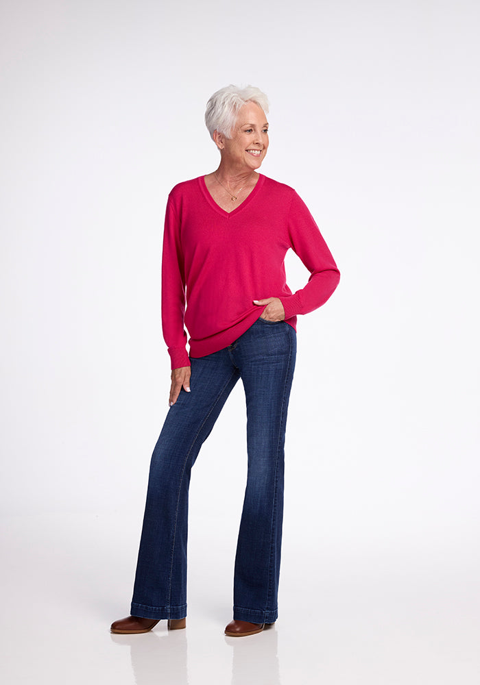 A smiling elderly person with short white hair stands confidently posing in a Woolx Francesca Sweater in Berry Burst and blue jeans, with one hand in their pocket. The background is plain white.