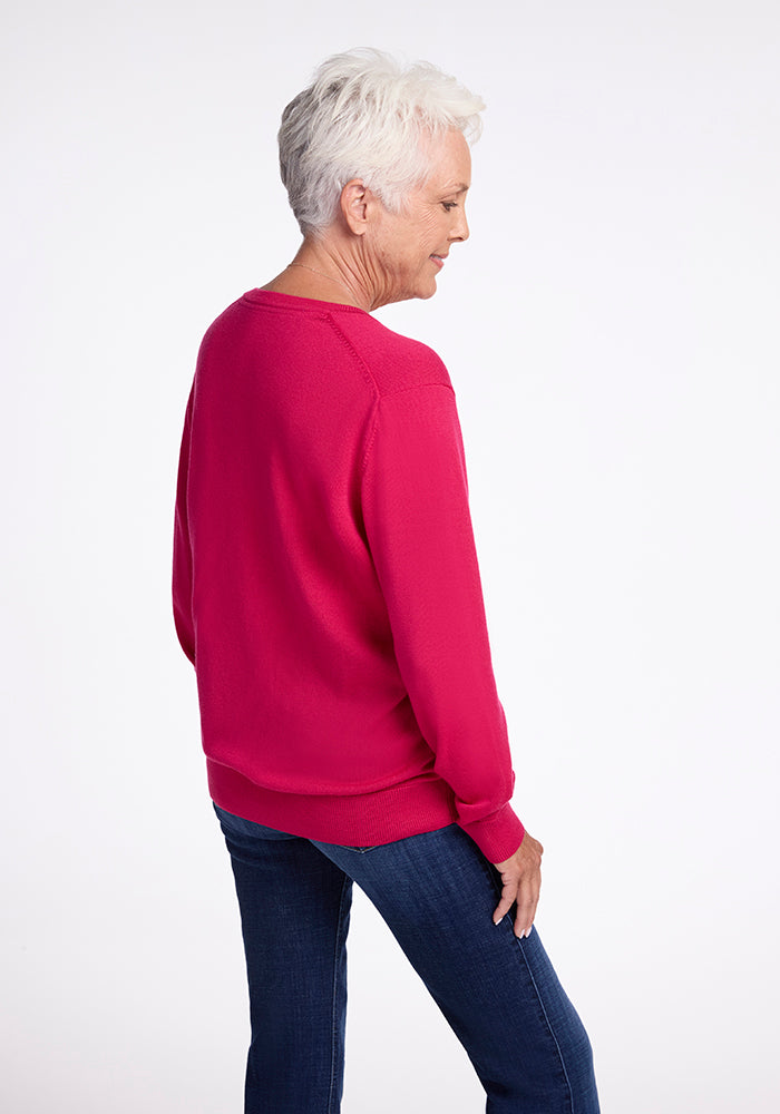 An older individual with short white hair is seen in a side profile, standing against a plain white backdrop. They are dressed in the elegant Francesca Sweater - Berry Burst from Woolx paired with dark blue jeans, looking down with a serene expression.