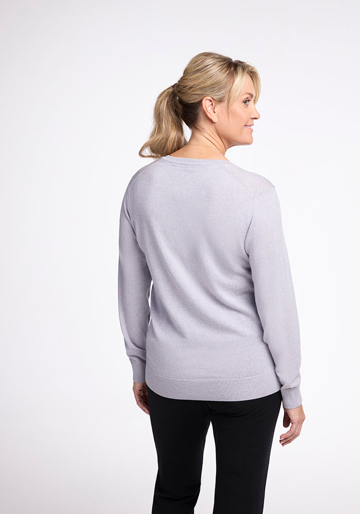 A woman with blonde hair tied in a ponytail is shown from the side, looking to her right. She is wearing the Woolx Francesca Sweater in Mist with black pants. The background is plain white.