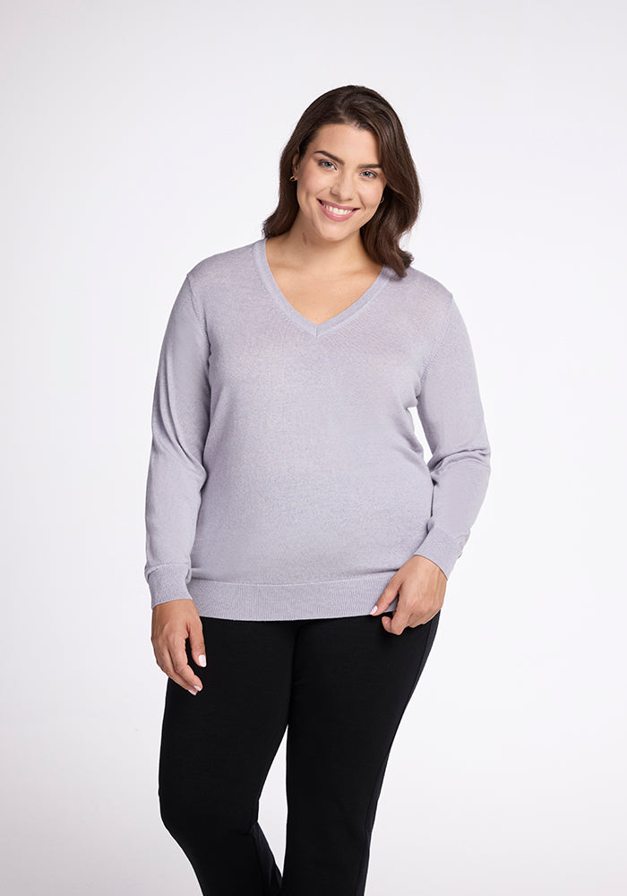 A woman with long brown hair is smiling while wearing the Francesca Sweater in Mist by Woolx and black pants. She stands against a plain white background.