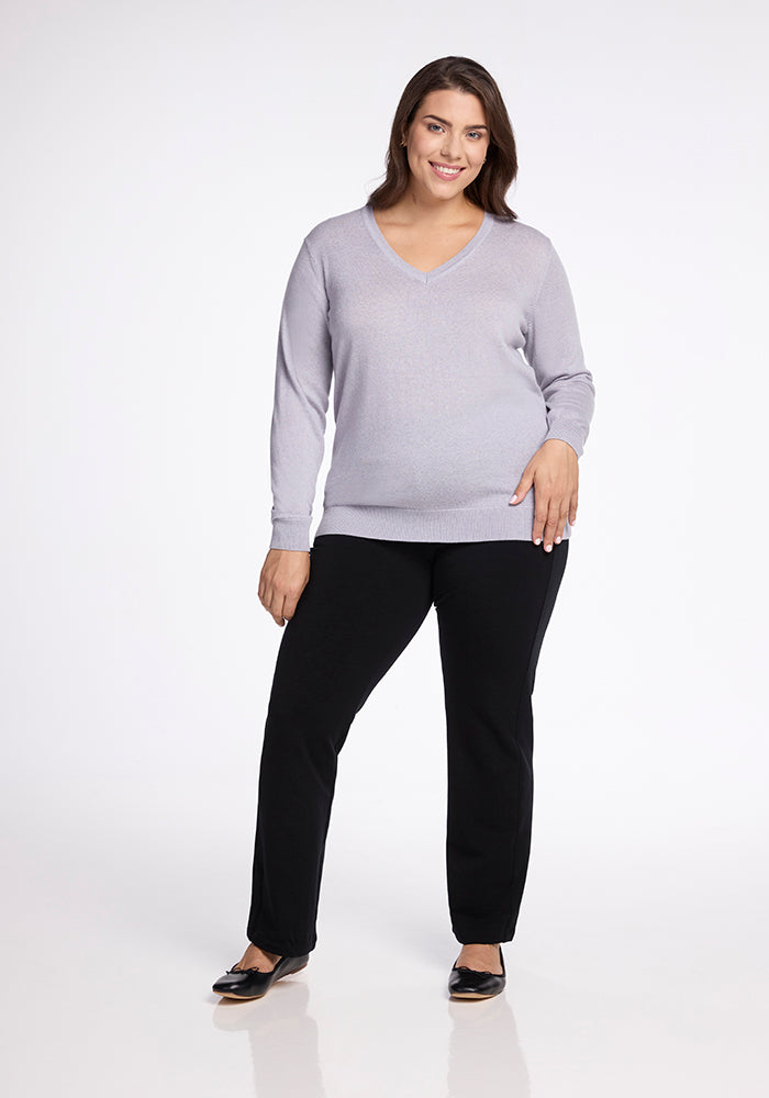 A smiling person is standing against a white background, wearing the Francesca Sweater in Mist by Woolx, paired with black pants and black shoes. They have one hand resting on their hip and the other hanging naturally by their side.