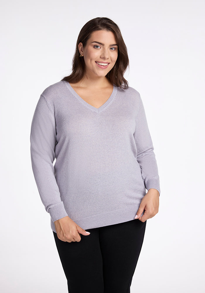 A person with shoulder-length dark brown hair is smiling while wearing the Francesca Sweater in Mist from Woolx and black pants. The background is plain white. 