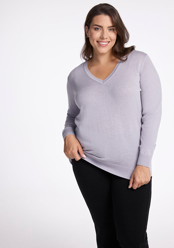 A person with long brown hair is smiling and looking at the camera, wearing the Francesca Sweater in Mist by Woolx paired with black pants. The background is plain white.