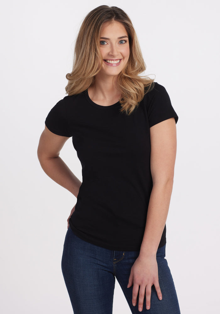 A person with long hair is smiling and standing with one hand on their hip. They are wearing the Woolx Addie Short Sleeve Crew in black along with blue jeans, set against a plain white background, highlighting the style and comfort of Australian Merino Wool that maintains freshness throughout the day. 