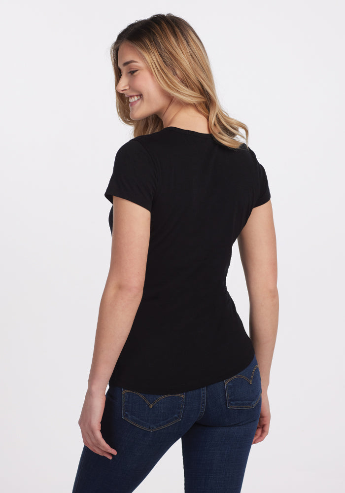 A woman with long hair, wearing a fitted Addie Short Sleeve Crew in black by Woolx and dark jeans, is smiling and looking to the side. She stands against a plain white background, her hands relaxed by her sides.