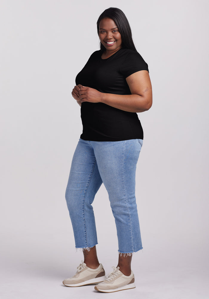 A person with long hair smiles while standing on a plain white background, wearing a black Woolx Addie Short Sleeve Crew tee, light blue jeans with frayed hems, and beige sneakers. The Merino wool tee is known for being odor-free.